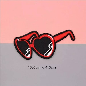 May include: Embroidered patch of heart-shaped sunglasses with red frames and black lenses. The patch measures 4.2 inches wide by 1.8 inches high. The sunglasses are set against a pink and gray background.