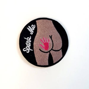 May include: A round, black patch with the phrase "Spank Me" embroidered in white. The patch features a stylized illustration of a rear with a pink handprint. The background is white.