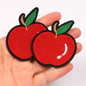 May include: Two embroidered apple patches. Each patch features a vibrant red apple with a black border, a green leaf, and a brown stem. One apple has a white curved accent. These patches are ideal for adding a touch of fruit-themed flair to clothing or accessories.