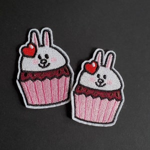 May include: Two embroidered iron-on patches featuring a white bunny rabbit with red hearts for eyes, inside a pink and brown cupcake.