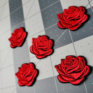 May include: Five red embroidered rose patches on a gray background. The roses are all the same size and shape, and they have a black outline.