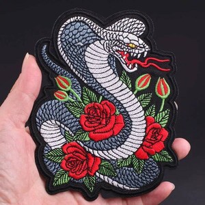 May include: Embroidered iron-on patch featuring a white cobra with red eyes, wrapped around red roses with green leaves on a black background.