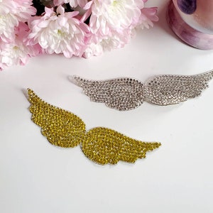 Puede incluir: Dos pares de alas de ángel hechas de diamantes de imitación. Un par es dorado y el otro es plateado.