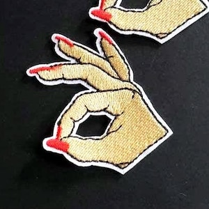 以下が含まれることがあります： OKサインを作るハンドジェスチャーが刺繍された2つのワッペン。手はベージュの糸で細部までステッチされ、爪は鮮やかな赤の糸で強調されています。各ワッペンには白い縁取りが施されています。