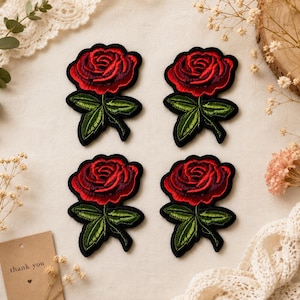 May include: Four embroidered rose patches. Each patch features a vibrant red rose with detailed petals, green leaves, and a black border. The patches are arranged on a cream-colored surface, suggesting a craft or sewing project.