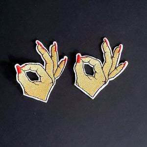 May include: Two embroidered iron-on patches of hands making the OK sign with red nail polish.