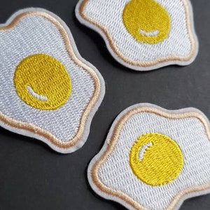 May include: Three embroidered iron-on patches shaped like fried eggs with yellow yolks. The patches are white with a brown outline.