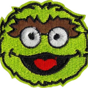 May include: Embroidered patch featuring a cartoon character with green fur, brown eyebrows, and large white eyes. The character has a wide, smiling mouth with a red tongue and black outline. The patch is outlined in black.
