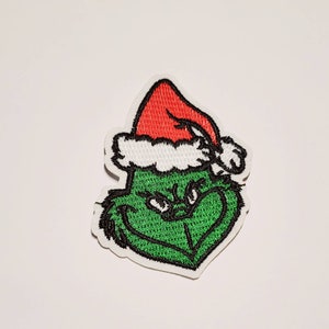 Puede incluir: Parche bordado con un personaje verde con una sonrisa traviesa, usando un gorro de Papá Noel rojo con ribete blanco. El parche tiene un borde blanco, ideal para ropa o accesorios.