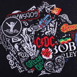 Puede incluir: Una colección de parches bordados de colores para planchar con logotipos de bandas y otros diseños, incluyendo "Green Day", "AC/DC", "Bob Marley", "Bowie" y "Godsmack".