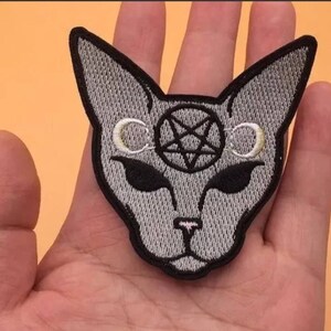 以下が含まれることがあります： 猫の顔のグレーの刺繍アイロン接着パッチ。目の上に五芒星と三日月が2つある。