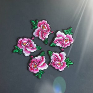 May include: Five embroidered floral patches. Each patch features a pink and white peony with green leaves and a yellow center. The patches are arranged in a circular pattern against a dark gray background.