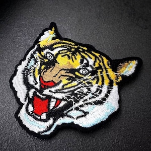 Könnte beinhalten: Nahaufnahme eines gestickten Tigergesichts-Patches. Der Tiger hat gelbe und schwarze Streifen, eine weiße Schnauze und einen roten, offenen Mund. Der Patch ist perfekt, um Kleidung, Taschen oder anderen Accessoires einen Hauch von Wildheit zu verleihen.