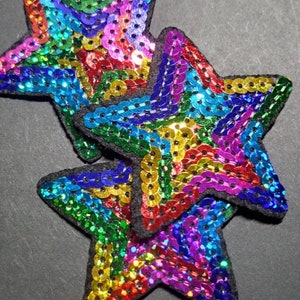 May include: Two rainbow sequined star-shaped patches. The stars are made of iridescent sequins in shades of red, orange, yellow, green, blue, indigo, and violet.