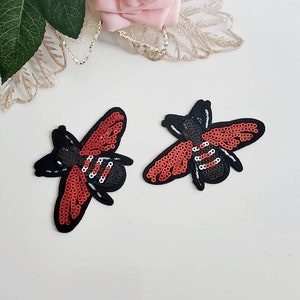 Può includere: Due toppe termoadesive a forma di ape rossa e nera con dettagli di paillettes. Le api hanno un corpo nero, ali rosse e strisce bianche.