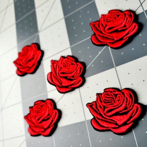 May include: Five red rose patches with black outlines are arranged on a gray and white grid surface. The embroidered roses have detailed petals, creating a realistic floral design. These patches are suitable for fabric embellishment.