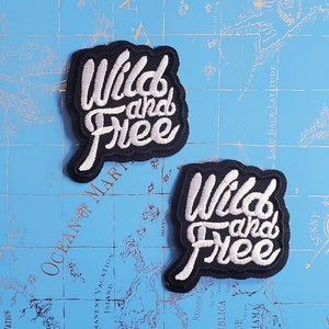 Può includere: Due toppe ricamate in bianco e nero con il testo "Wild and Free" in un font corsivo. Le toppe sono su uno sfondo blu.