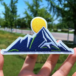 Könnte beinhalten: Ein gestickter Aufnäher, der eine blaue und weiße Bergkette mit einer gelben Sonne darüber zeigt. Der Aufnäher hat einen weißen Hintergrund und grüne Akzente.