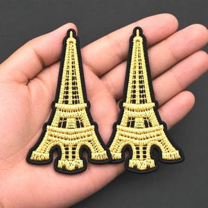 May include: Two embroidered patches shaped like the Eiffel Tower. Each patch features metallic gold thread detailing the structure of the tower against a solid black background, creating a distinct contrast between the gold and black colors.