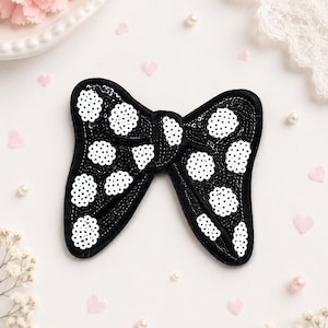 May include: A black bow-shaped applique featuring a pattern of white circular sequin clusters. The bow is constructed with a black base and is densely covered in black sequins, with white sequins arranged in distinct polka dot formations.