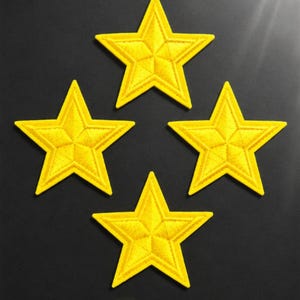 Könnte beinhalten: Vier leuchtend gelbe sternförmige Aufnäher, die in einem Rautenmuster angeordnet sind. Jeder gestickte Stern weist ein zentrales Fünfeck-Design mit nach außen verlaufenden Stepplinien auf, was eine texturierte, geometrische Optik erzeugt.