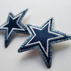 Puede incluir: Dos parches en forma de estrella azul marino con detalles blancos y azul brillante. Las estrellas tienen un diseño de cinco puntas con un contorno blanco. Estos parches son probablemente para uso decorativo en ropa o accesorios.
