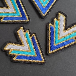 May include: Four embroidered arrow patches in blue, turquoise, white, and gold. The arrows are pointing downwards.
