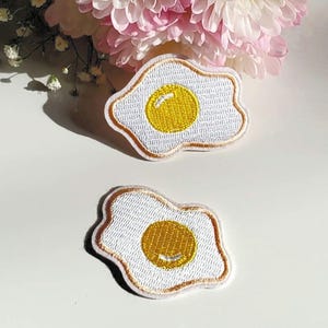 Puede incluir: Dos parches bordados con forma de huevos fritos. Cada parche tiene un fondo blanco, una yema amarilla y un borde marrón. Los parches están sobre una superficie blanca con flores rosas y blancas en el fondo.