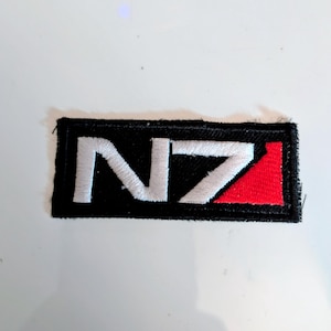 May include: Embroidered patch featuring the letters "N7" in white, set against a black rectangle with a red triangle on the right side. The patch is likely for clothing or accessories.