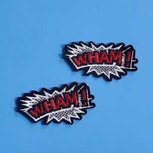 以下が含まれることがあります： 青い背景に白い輪郭のある赤い刺繍パッチが2つ。各パッチには、コミックスタイルのフォントで「WHAM！」という単語が書かれています。