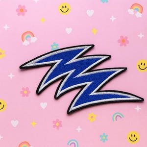 Puede incluir: Parche bordado de rayo azul con ribete blanco y negro. El parche tiene forma de zigzag. El fondo es rosa con varios elementos decorativos coloridos, incluyendo flores, arcoíris, corazones y caras sonrientes.