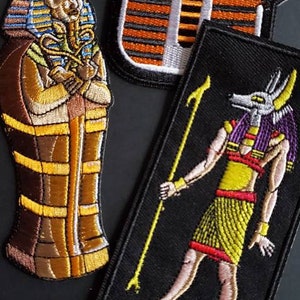 May include: Three embroidered iron-on patches featuring Egyptian themes. The first patch is a yellow and orange pharaoh's head with a red headdress. The second patch is a brown and gold sarcophagus with a blue and gold pharaoh inside. The third patch is a black background with a yellow and gold Anubis figure holding a staff.