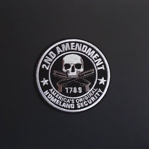Könnte beinhalten: Ein gestickter schwarzer und weißer Aufnäher mit einem Totenkopf und gekreuzten Knochen, dem Text "2nd Amendment" und dem Text "America's Original Homeland Security" mit dem Jahr "1789" in der Mitte.
