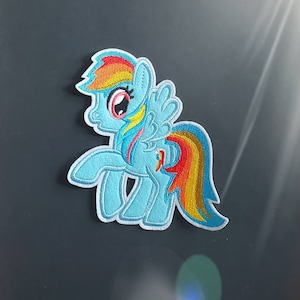 May include: A blue and yellow embroidered patch of a cartoon pony with a rainbow mane and tail. The pony has a pink nose and a blue eye.
