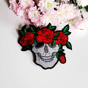 May include: White embroidered skull with a red rose crown. The skull has a toothy grin and is surrounded by green leaves.
