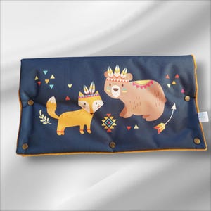 Könnte beinhalten: Ein blaues Stoffkissen mit gelbem Rand zeigt einen Fuchs und einen Bären, die farbenfrohe Kopfbedeckungen tragen. Der Hintergrund ist dunkelblau mit einem geometrischen Muster.