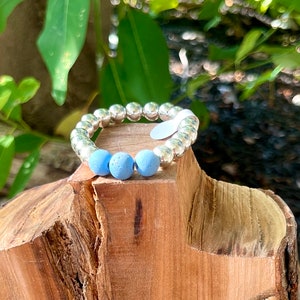May include: A beaded bracelet with silver-colored beads and three light blue beads. The bracelet is displayed on a wooden stand, with green foliage in the background.