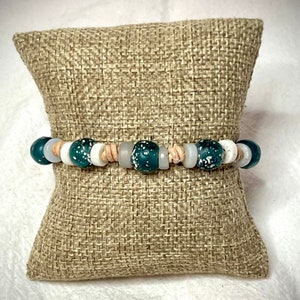 May include: A green and white beaded bracelet with brown spacers. The bracelet is displayed on a brown burlap cushion.