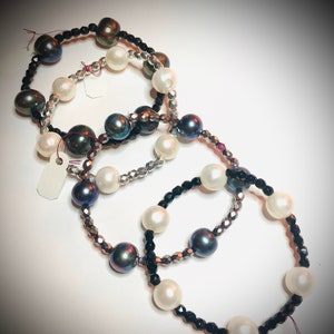 May include: Three pearl bracelets with black, silver, and iridescent beads. The bracelets are layered on a white surface.