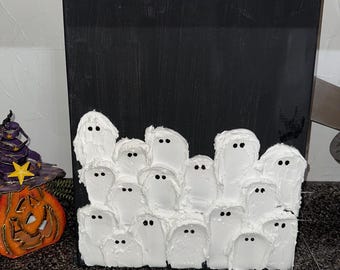 Puede incluir: Un lienzo negro con un diseño de fantasmas blancos texturizados. Los fantasmas tienen ojos de puntos negros y están dispuestos en un grupo. Una calabaza de Halloween y un sombrero de bruja son visibles en el fondo.