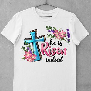 May include: White t-shirt with a blue cross with a leopard print design, pink flowers, and the text "He is Risen indeed".