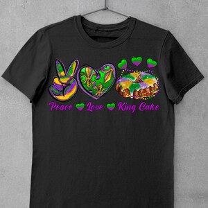 May include: Black t-shirt with a peace sign, heart, and king cake graphic. The graphic is in purple, green, and gold colors. The text "Peace Love King Cake" is printed below the graphic.
