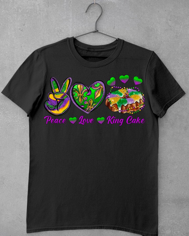 May include: Black t-shirt with a peace sign, heart, and king cake graphic. The graphic is in purple, green, and gold colors. The text "Peace Love King Cake" is printed below the graphic.