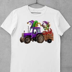 Pu&ograve; includere: Una maglietta bianca con un trattore viola che tira un carro pieno di gamberi di fiume. Il trattore ha un cappello da giullare in cima e una maschera di Mardi Gras verde e oro &egrave; nel carro.