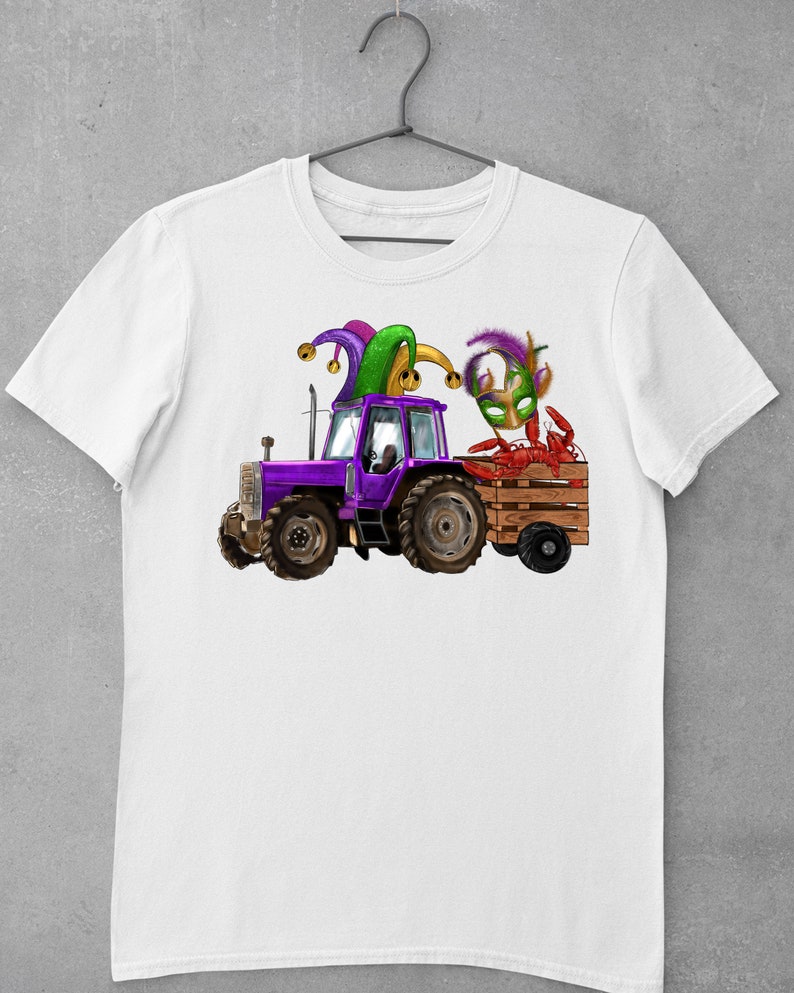 Pu&ograve; includere: Una maglietta bianca con un trattore viola che tira un carro pieno di gamberi di fiume. Il trattore ha un cappello da giullare in cima e una maschera di Mardi Gras verde e oro &egrave; nel carro.