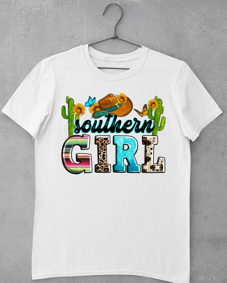 Pode incluir: Camiseta branca com um design gr&aacute;fico de um chap&eacute;u de cowboy, girass&oacute;is, borboletas e cactos. O texto "southern girl" est&aacute; escrito em letras coloridas.
