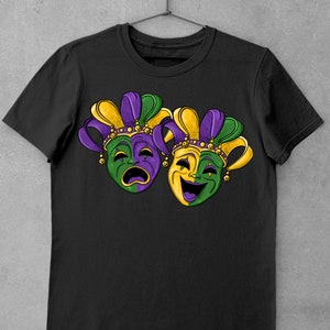 May include: Black t-shirt with a graphic of two Mardi Gras masks. One mask is green and purple with a sad expression, and the other is yellow and green with a happy expression.