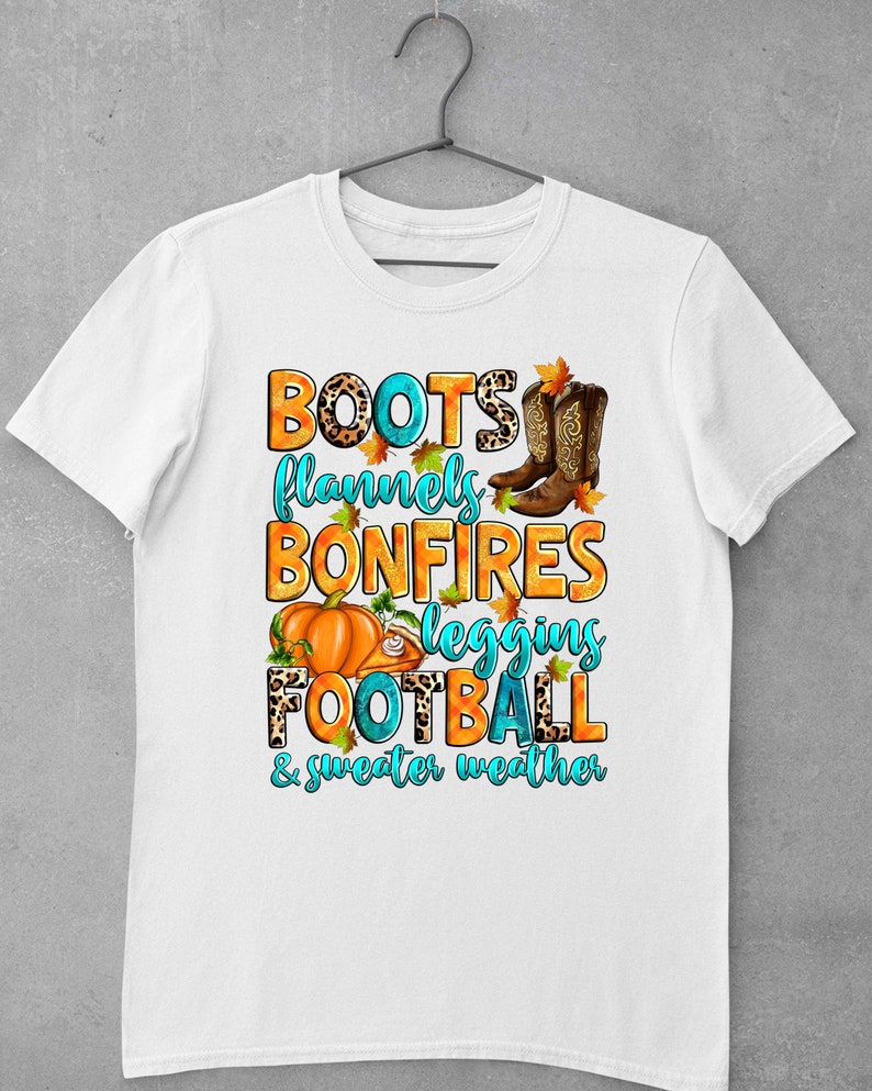 May include: White t-shirt with a graphic design featuring the text "Boots, flannels, bonfires, leggings, football, & sweater weather" with images of boots, a pumpkin, a football, and autumn leaves.