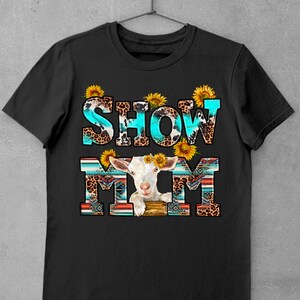 May include: Black t-shirt with the text "SHOW MOM" in turquoise and white letters with a white goat with sunflowers on its head in the middle of the text. The letters are decorated with a leopard print and turquoise and brown geometric patterns.