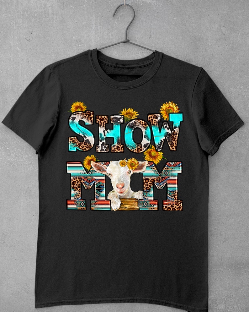 May include: Black t-shirt with the text "SHOW MOM" in turquoise and white letters with a white goat with sunflowers on its head in the middle of the text. The letters are decorated with a leopard print and turquoise and brown geometric patterns.